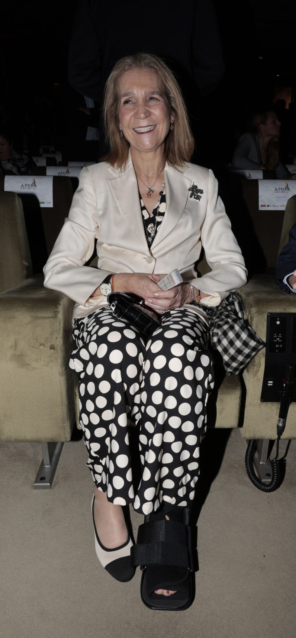 la infanta elena sonrie, con un mono de lunares en blanco y negro y una bota walker en su pie izquierdo