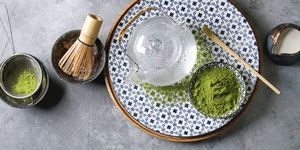 ingredients for making matcha ice drink green tea matcha powder in ceramic bowl, traditional bamboo spoon, whisk on plate, glass teapot, ice cubes over grey texture background flat lay, space