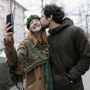 a man kissing his girlfriend on the cheek as she takes a selfie of the two of them while out for a walk in the city together