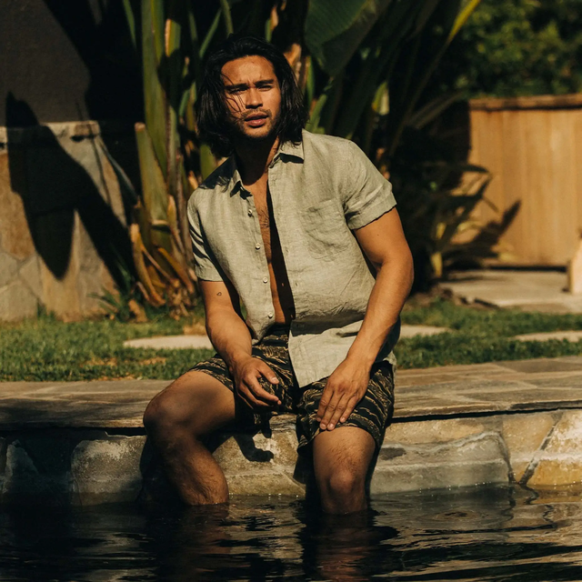 man sitting on edge of pool with legs in water wearing taylor stitch shirt and swim trunks