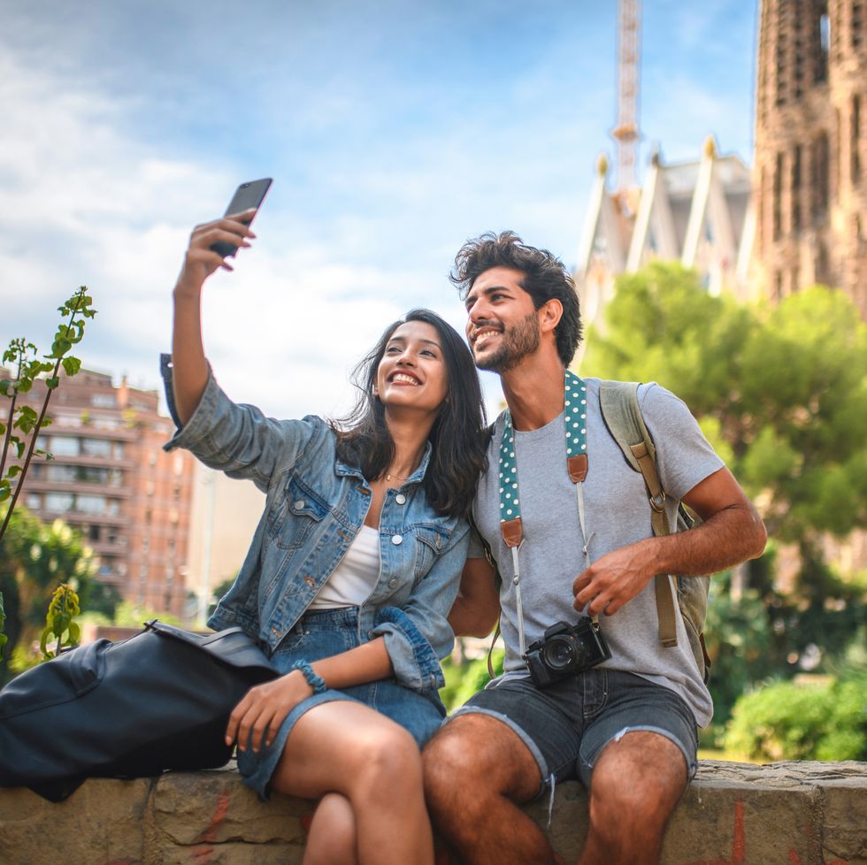 male and female travelers sitting on wall in public park near sagrada familia in barcelona and taking selfie on sunny summer day