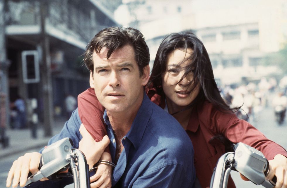 pierce brosnan wearing a blue shirt, and michelle yeoh wearing a red shirt, sitting on a motorcycle in a city road