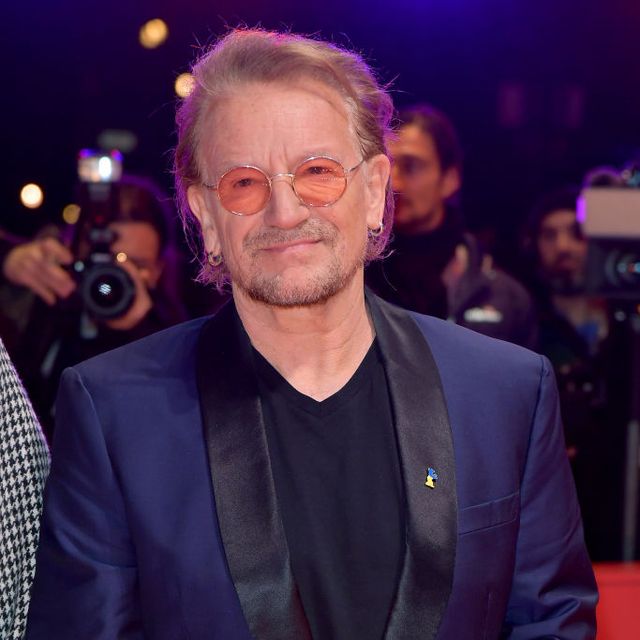 bono smiles and looks past the camera, he stands on a red carpet with photographers behind him, he wears a blue and black suit jacket with a black shirt, orange circular glasses, and earrings