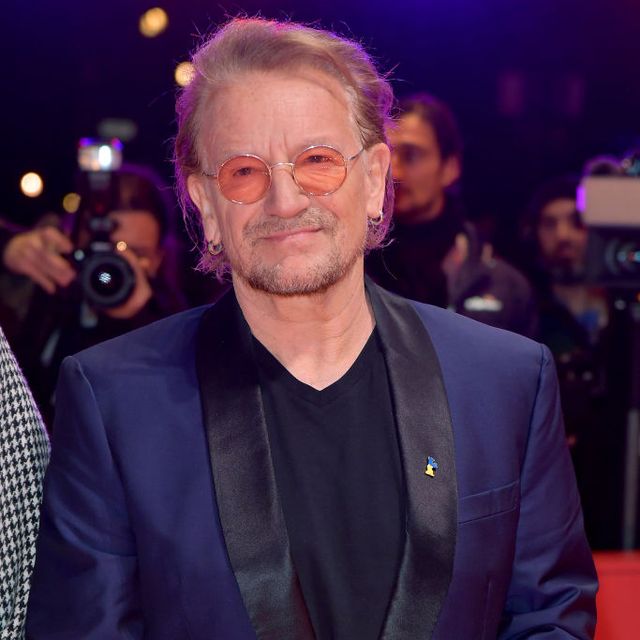 bono smiles and looks past the camera, he stands on a red carpet with photographers behind him, he wears a blue and black suit jacket with a black shirt, orange circular glasses, and earrings