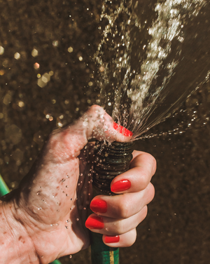 a woman wearing red nail polish holds her thumb over a squirting garden hose