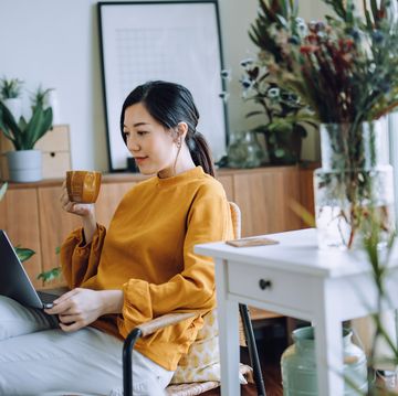 young asian woman sitting on armchair using laptop while relaxing in living room at home, surrounded by green houseplants lifestyle and technology