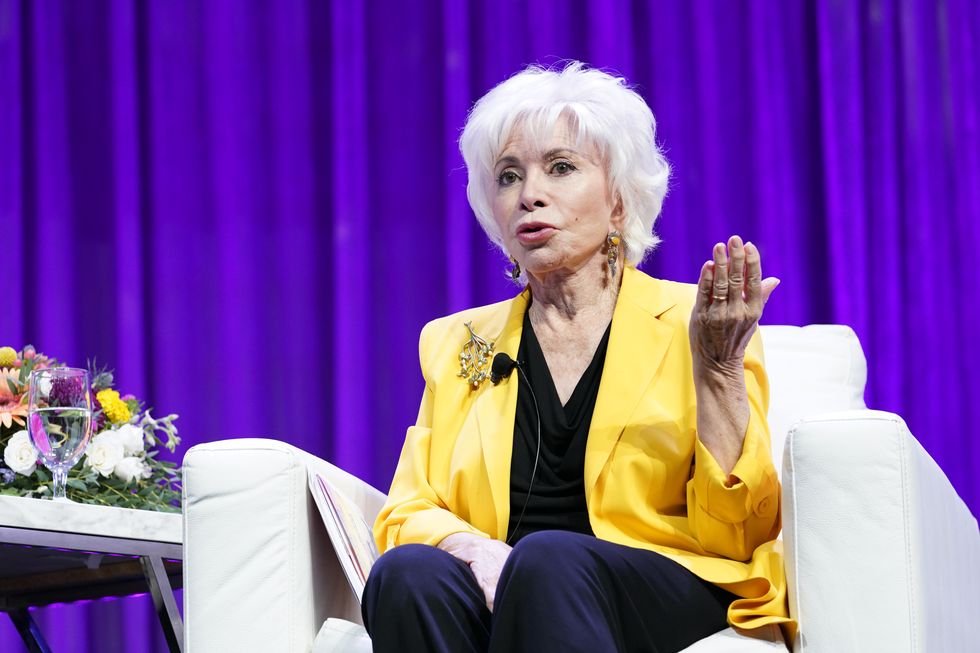 isabel allende sits in an armchair and talks with one hand raised, she wears a yellow suit jacket, black shirt and dark pants