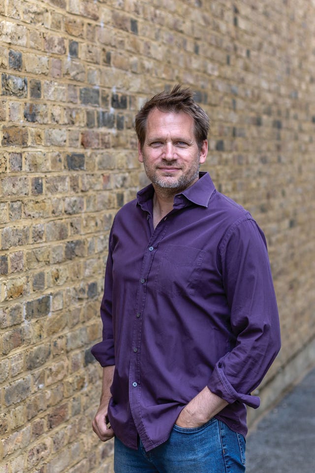 portrait of john freeman standing in front of a brick wall, wearing a purple button down shirt, hands in pockets, gazing into the camera.
