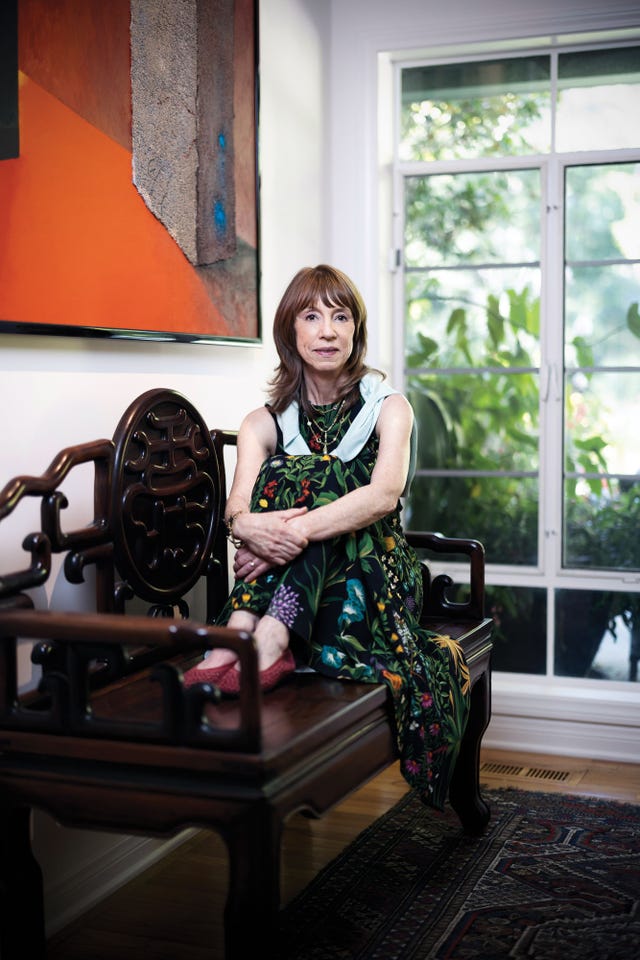 lisa see seated on a carved wooden bench in her home, wearing a floral dress, surrounded by bright sunlight and greenery through a window behind her.
