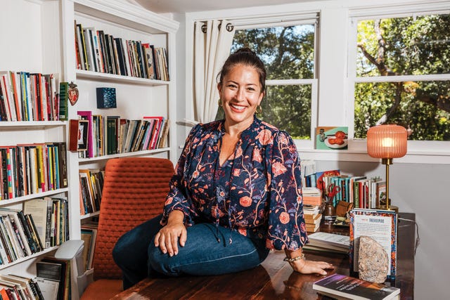 poet ada limón sits on a desk in front of bookshelves in her california home, smiling in a floral blouse, sunlight glowing through the window behind her.