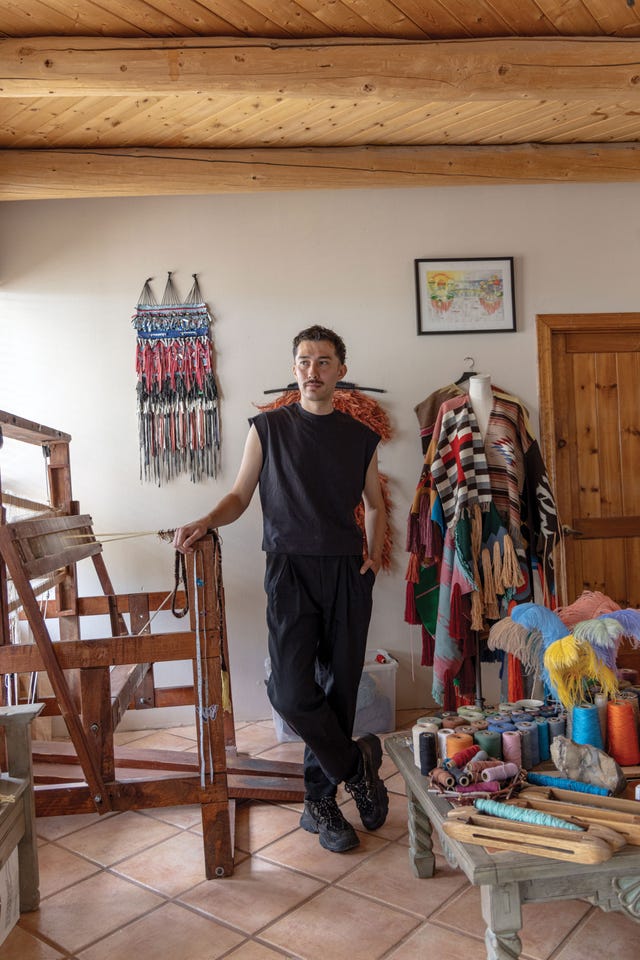 josh tafoya stands in his home weaving studio in ranchos de taos, surrounded by wool spools, looms, and woven garments in bright natural dyes.