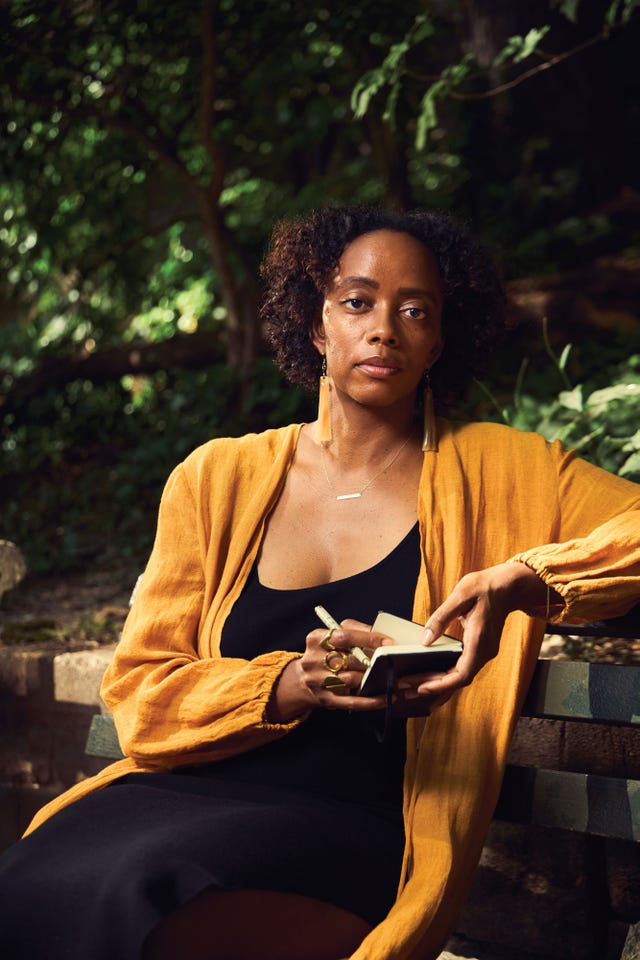angela flournoy seated on a park bench wearing a mustard shawl, holding a small notebook and pen, surrounded by trees and dappled light.