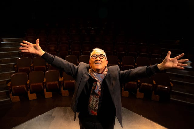 playwright philip kan gotanda stands center stage with outstretched arms inside an empty theater, smiling in a dark jacket and scarf.