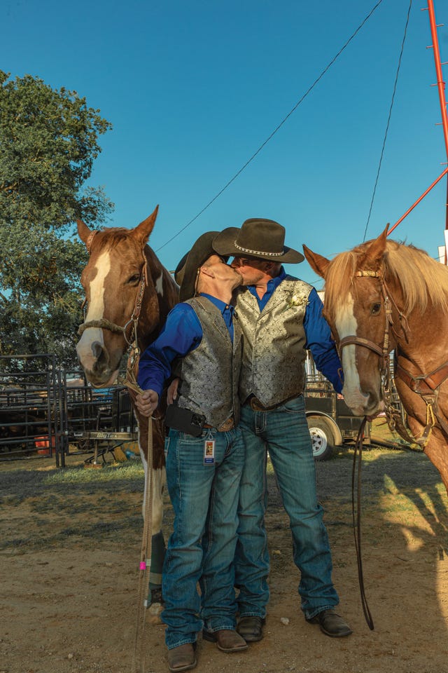 two cowboys in matching vests and blue shirts share a kiss between their horses at the denton fairgrounds.