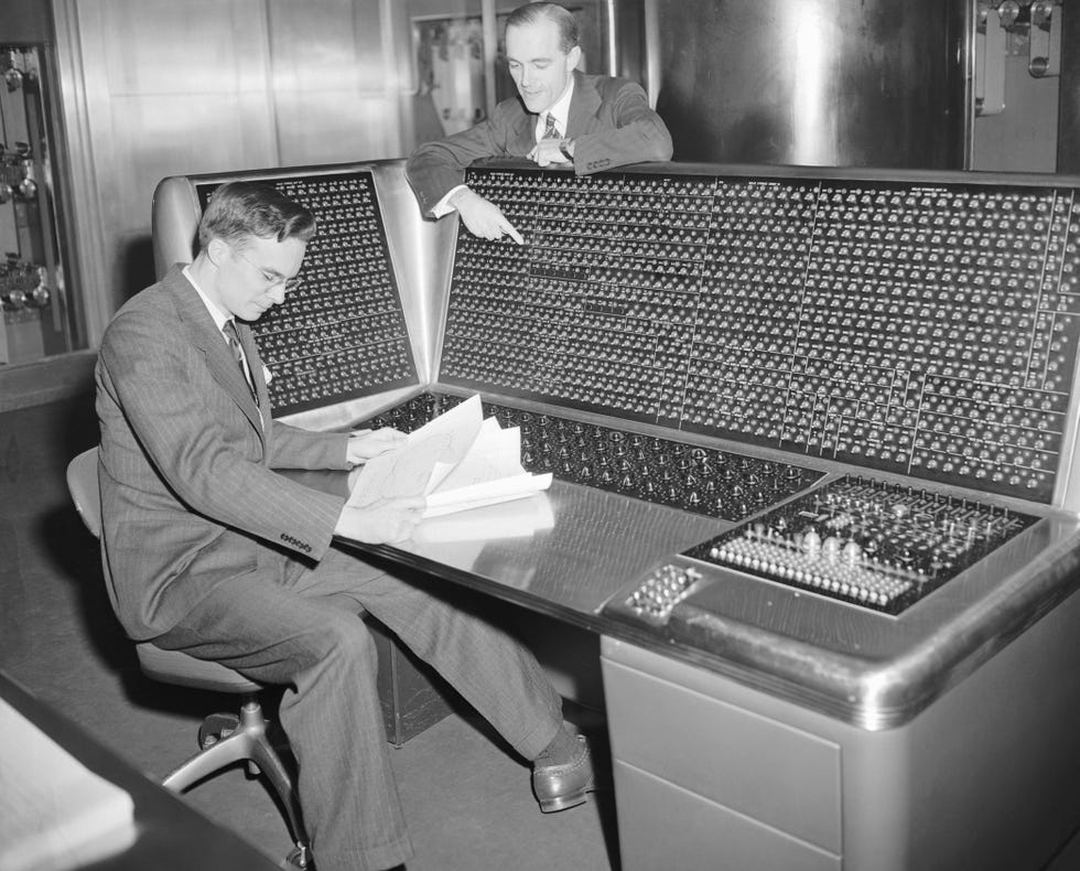 one man sits at a large supercomputer and looks at papers, another man points at the large supercomputer from the back