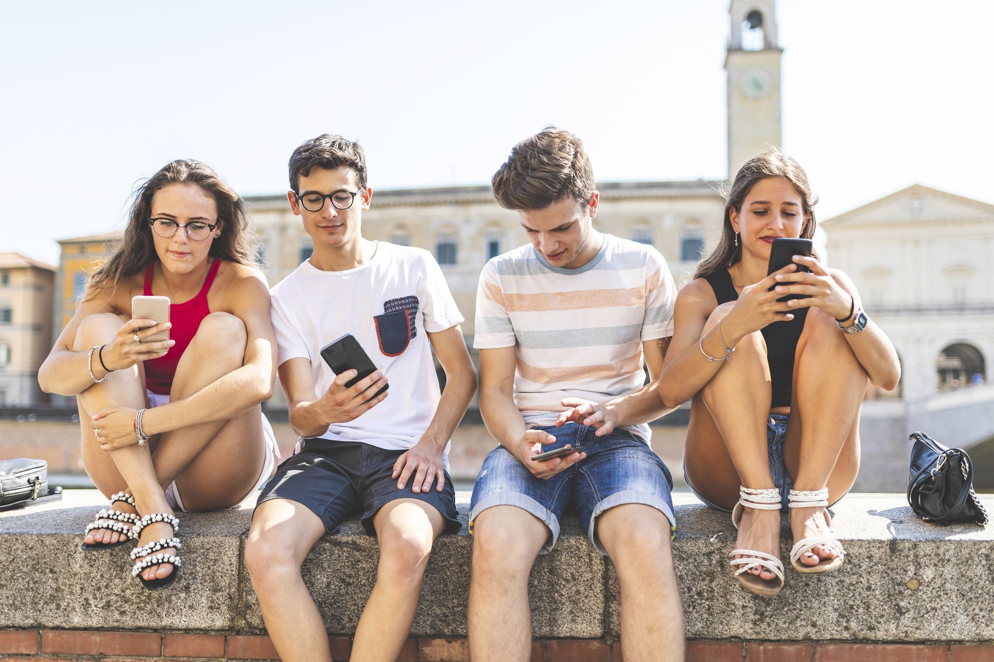 Italy, Pisa, group of four friends sitting together on a wall using cell phones