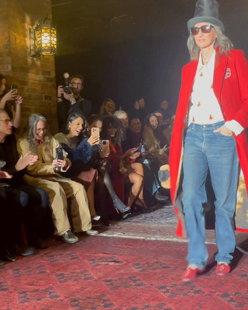 fashion model showcasing a vibrant red coat and casual jeans on a runway while an audience captures the moment