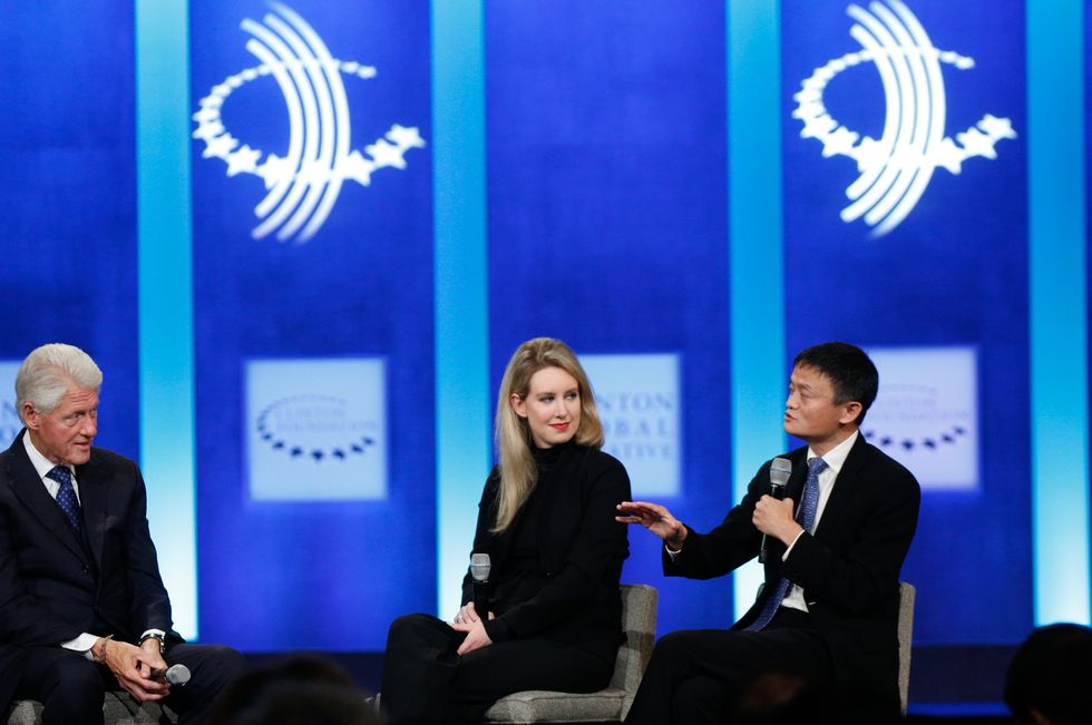 bill clinton, elizabeth holmes, and jack ma sit on a stage in chairs, ma is speaking into a microphone he holds as clinton and holmes watch