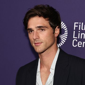 jacob elordi looks left of the camera while standing in front of a blue background, he wears a black suit jacket and stripped collared shirt unbuttoned at the neck