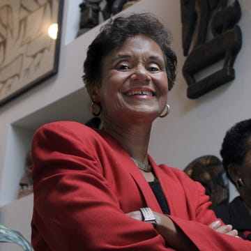 dr marilyn gaston smiles down at the camera while standing with her arms crossed, she wears a red suit jacket, gold hoop earrings, a gold necklace, and a gold bracelet, her short hair is styled with loose curls, and her makeup includes soft red lipstick, in the background on the right is dr gayle porter who also has short hair and is smiling while looking away from the camera