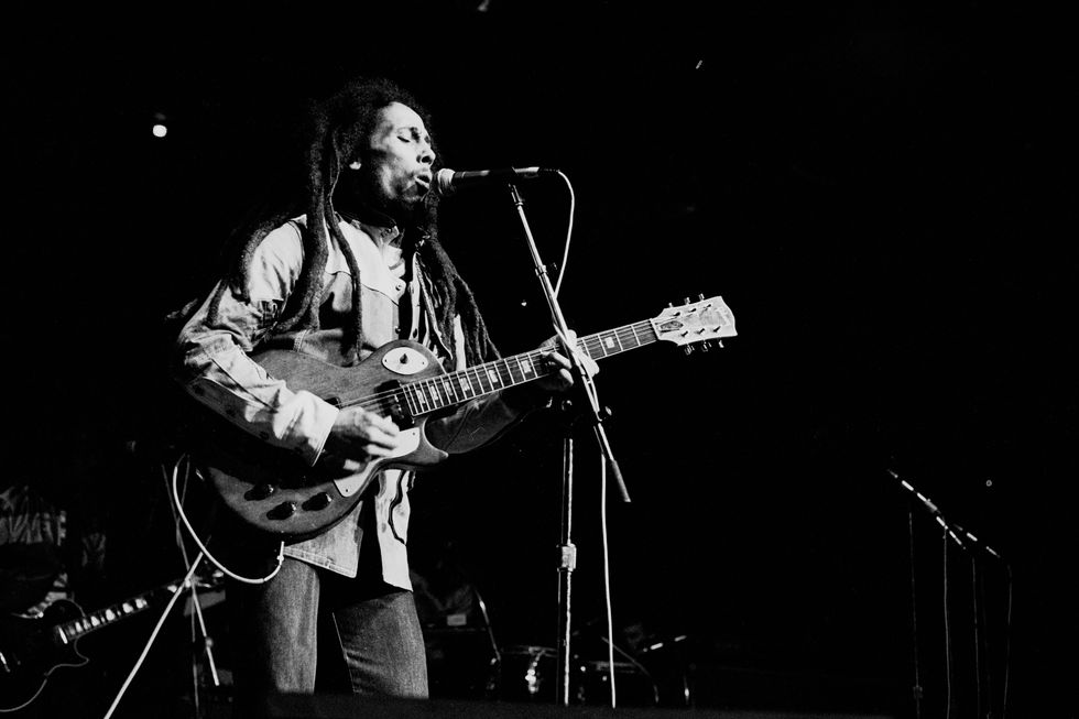 bob marley sings into a microphone and strums an electric guitar he wears on a strap, he has on a jean jacket and pants in this black and white photo