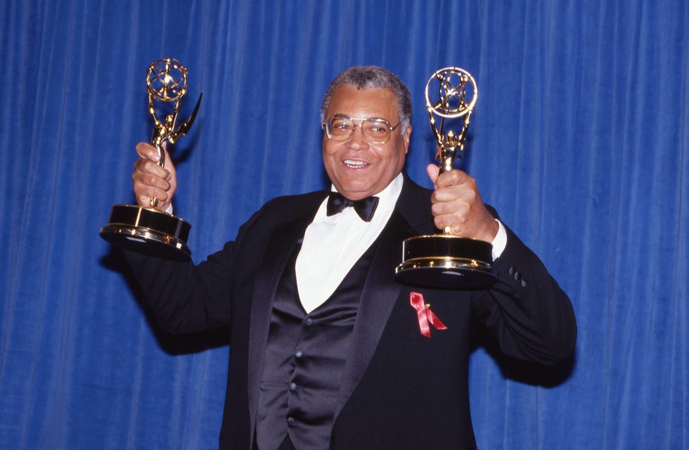 james earl jones stands and smiles as he raises two emmy trophies on either side of him, he wears a black tuxedo with a black vest