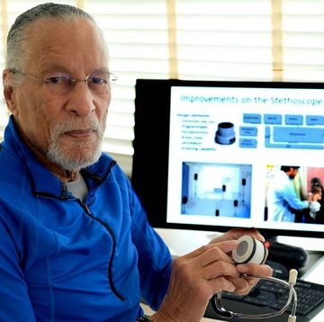 james west sits at a desk and looks at the camera, he holds a circular device in his hands, he wears a bright blue quarter zip top and glasses, behind him is a computer and window blinds