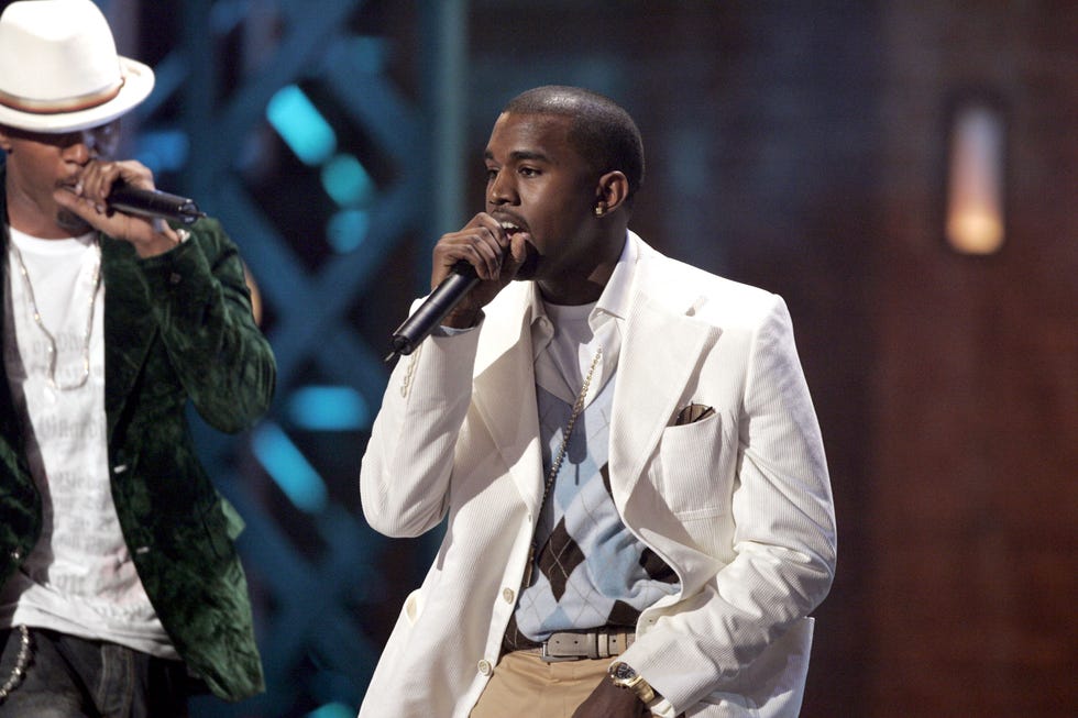 kanye west sings into a wireless microphone he holds in one hand as he stands on a stage, he wears a white suit jacket, argyle sweater, and khaki pants