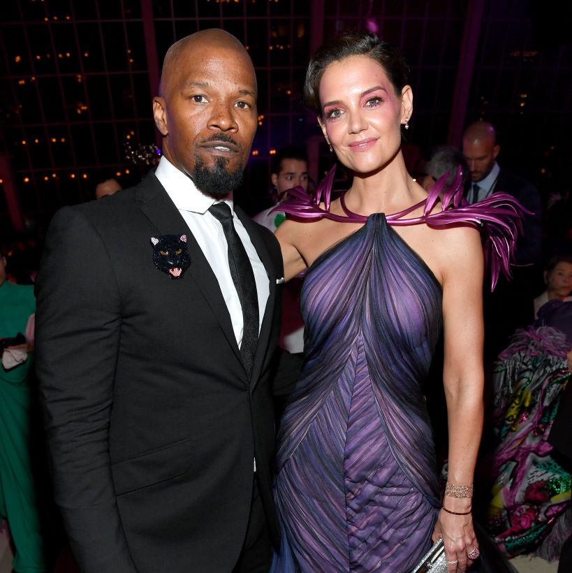jamie foxx and katie holmes stand and pose for a photo, he wears a black suit with a tie and a sparkly panther pin, she wears a purple gown and makeup