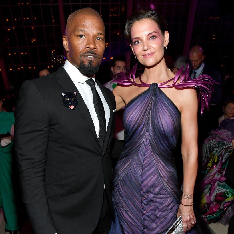 jamie foxx and katie holmes stand and pose for a photo, he wears a black suit with a tie and a sparkly panther pin, she wears a purple gown and makeup