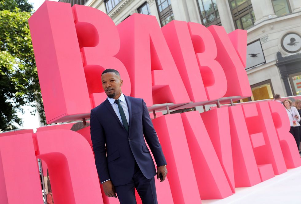 jamie foxx wearing a suit and tie, standing in front of a large display that spells the words baby driver