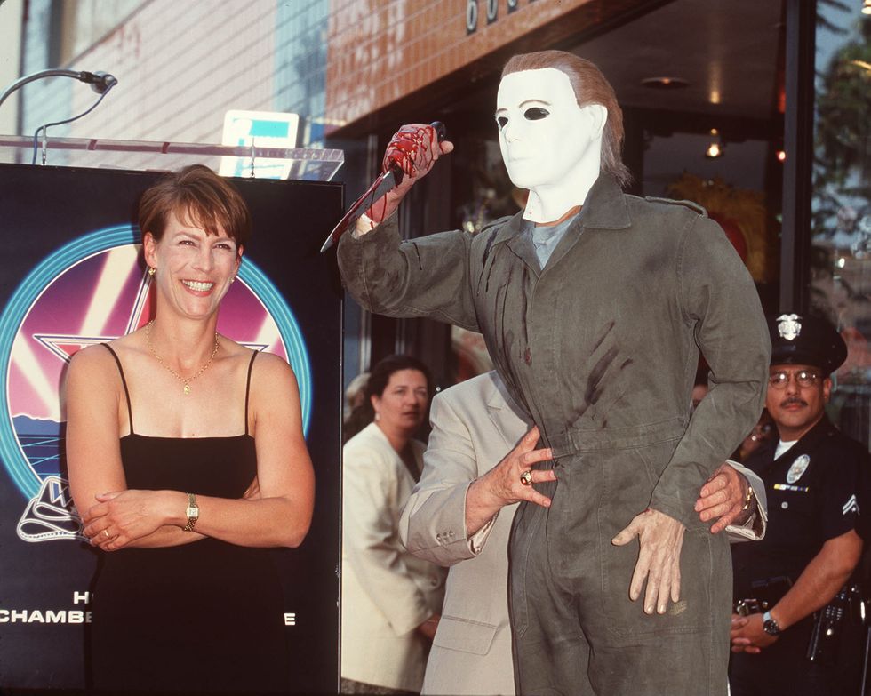 jamie lee curtis, wearing a black dress, crosses her arm and smiles as a man dressed as michael myers from the movie halloween holds a knife next to her
