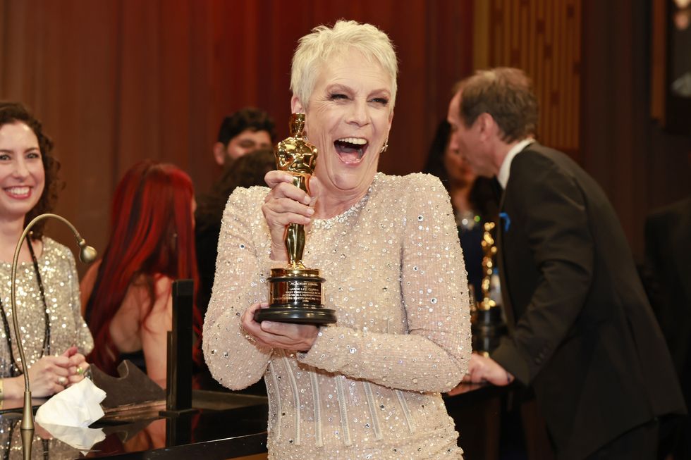 jamie lee curtis wearing a white sequined dress, smiling and holding an oscar statuette, with several people milling behind her