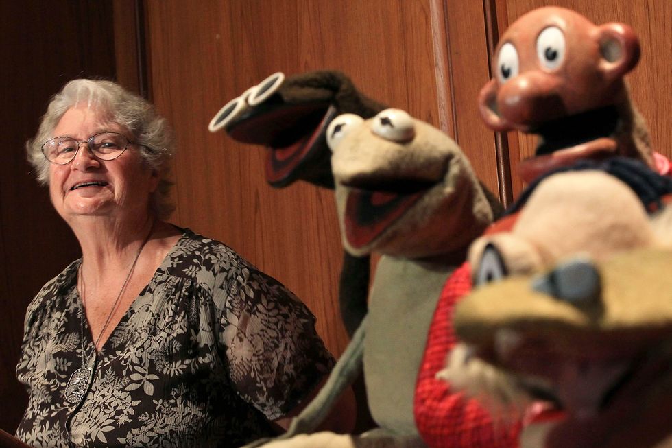 jane henson smiles while standing to the left of several muppets, she wears a patterned black and gray shirt