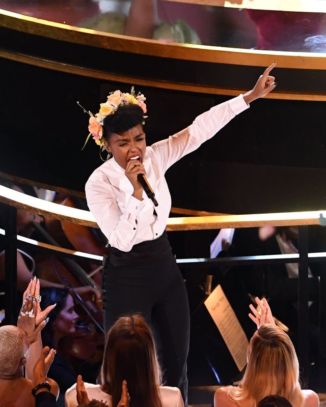 janelle monae, wearing a white tuxedo shirt and black pants, with flowers in her hair, stands in front of a stage and sings into a microphone, pointing her left hand in the air, while audience members in seats in front of her applaud