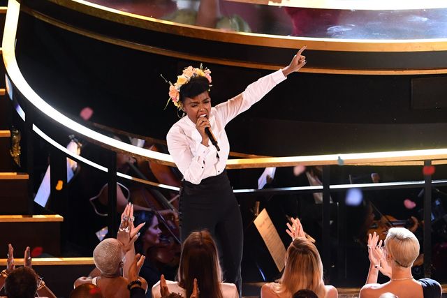 janelle monae, wearing a white tuxedo shirt and black pants, with flowers in her hair, stands in front of a stage and sings into a microphone, pointing her left hand in the air, while audience members in seats in front of her applaud