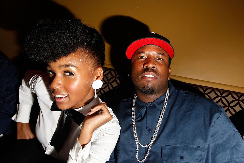janelle monáe, wearing a white tuxedo shirt, leans forward and smiles while sitting next to big boi, who wears a blue shirt, black and red hat, and a white necklace