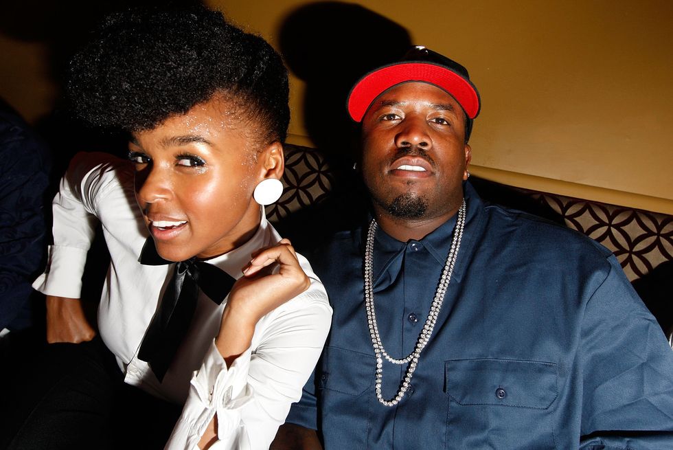 janelle monáe, wearing a white tuxedo shirt, leans forward and smiles while sitting next to big boi, who wears a blue shirt, black and red hat, and a white necklace