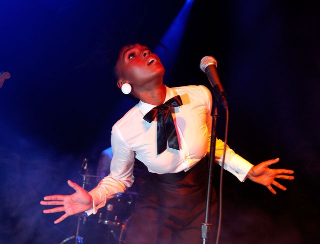 janelle monáe, wearing a white tuxedo shirt, black pants and a black ribbon tie, performs on a stage in front of a microphone, looking up at the sky with both arms out at her sides