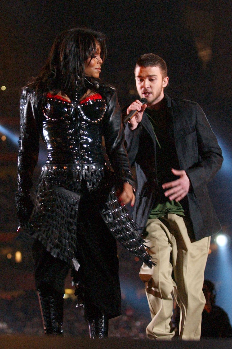 janet jackson and justin timberlake walk on a stage as he sings into a microphone, she wears a mostly black outfit, he wears a dark jacket over a green shirt and khaki pants
