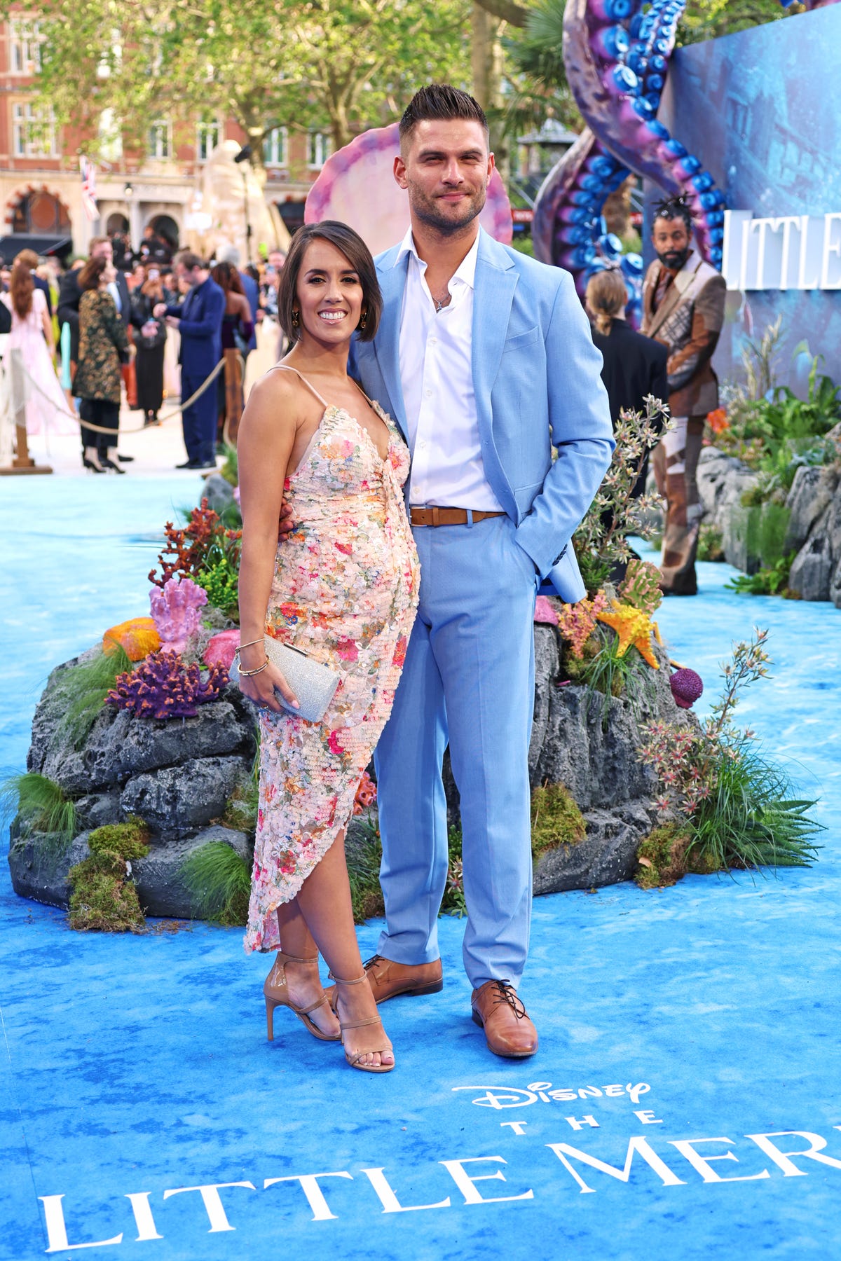 janette manrara wearing a floral spaghetti strap dress and holding a silver clutch while standing next to her husband aljaz skorjanec, who is wearing a white shirt and a light blue suit, standing together on the blue carpet at the uk premiere of the little mermaid in may 2023