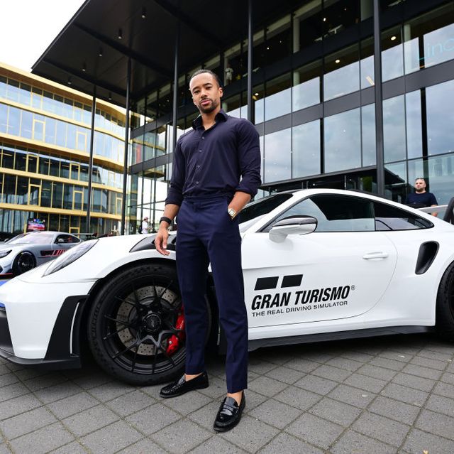 jann mardenborough poses in front of a white sports car with a gran turismo logo on the driver door, mardenborough wears a navy collared shirt, navy pants, and black loafers