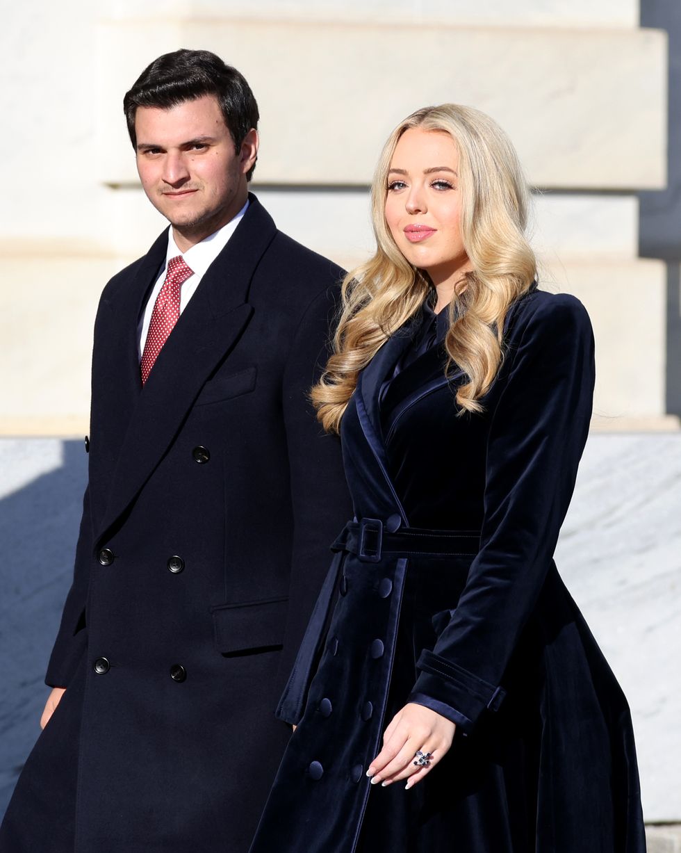 michael boulos and tiffany trump walk outside and look to the right, they both wear long coats