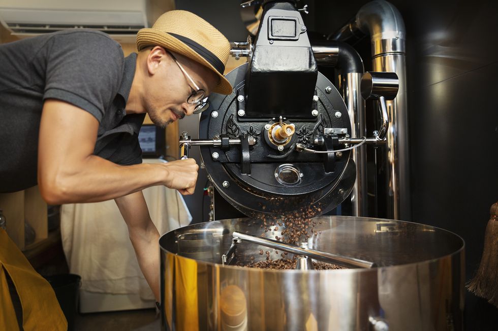 japanese man wearing hat and glasses standing in an eco cafe, operating coffee roaster machine