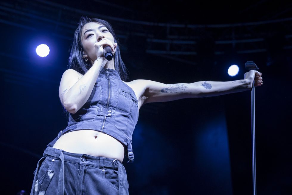 rina sawayama wearing black and performing on a stage, holding a microphone and leaning a hand on a microphone stand