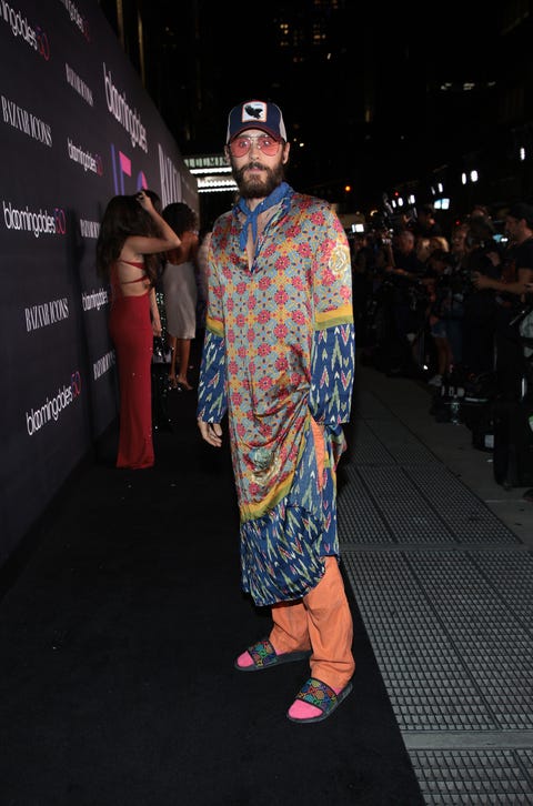 jared leto stands on the sidewalk outside bloomingdale's wearing a trucker hat, a multicolored caftan, orange pants, multicolored slides, pink socks, a blue neckerchief, and pink tinted sunglasses