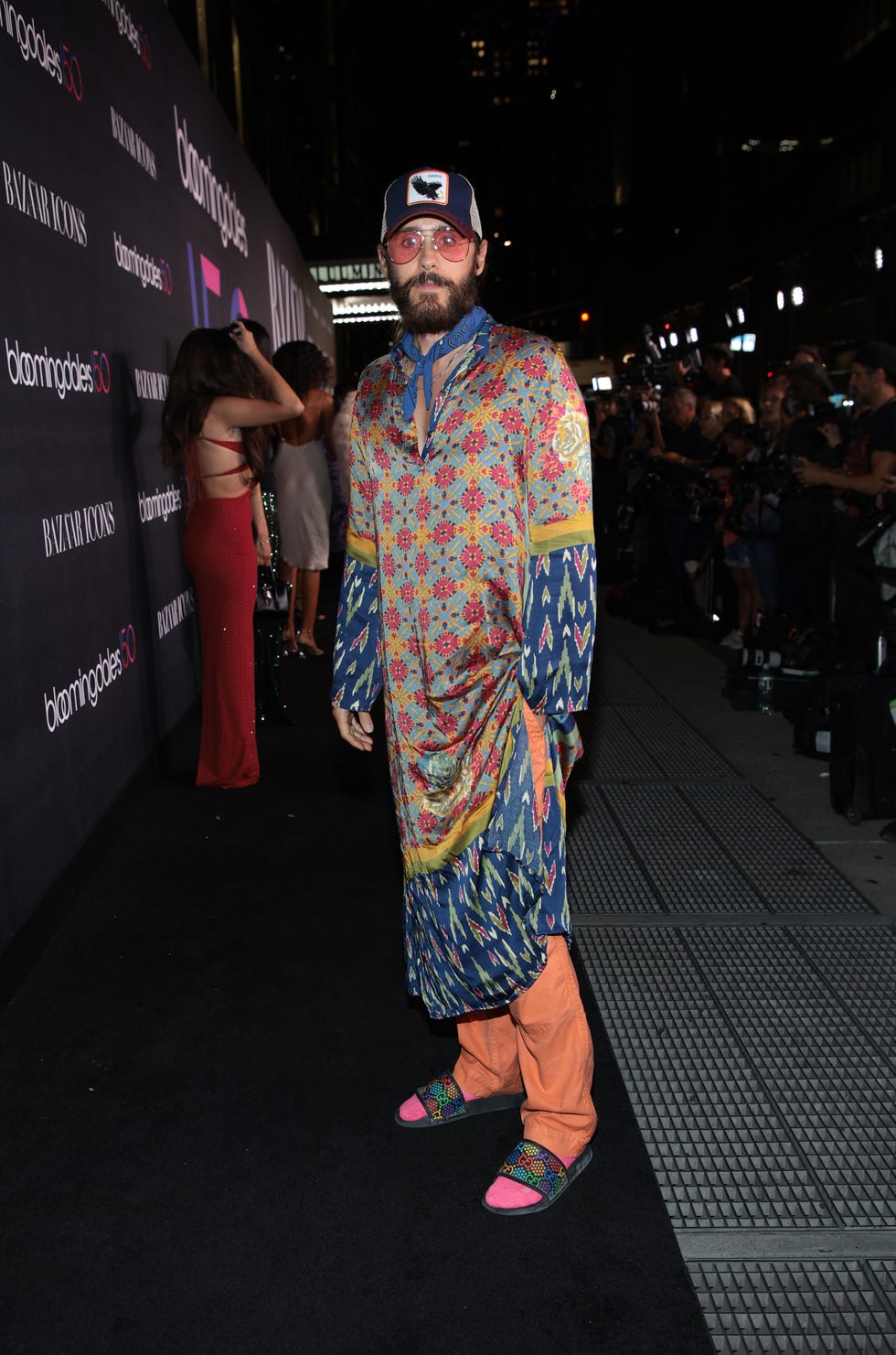 jared leto stands on the sidewalk outside bloomingdale's wearing a trucker hat, a multicolored caftan, orange pants, multicolored slides, pink socks, a blue neckerchief, and pink tinted sunglasses