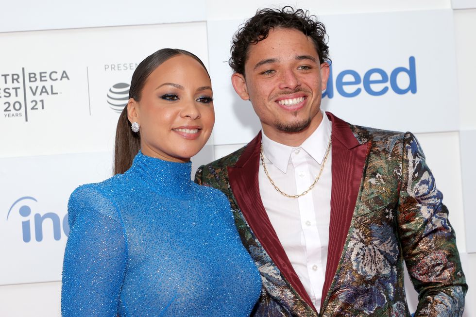 jasmine cephas jones, wearing a blue dress, and anthony ramos, wearing a patterned suit jacket and white shirt, smiling and looking off camera in front of a white wall with the tribeca film festival logo on it