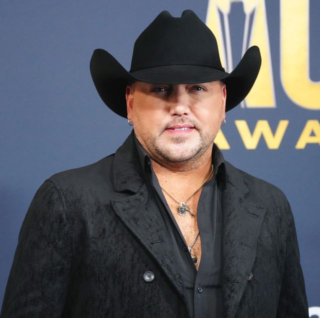 jason aldean looks at the camera with a small smile, he wears a black cowboy hat, a black jacket, black unbuttoned collared shirt, and two silver necklaces