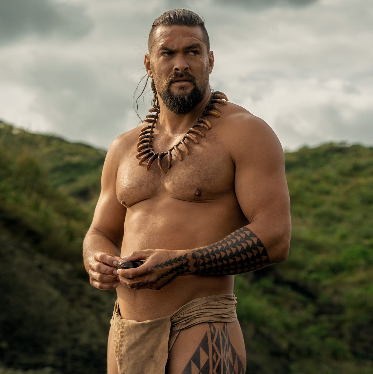Jason Momoa portrays Ka'iana in 'Chief of War' man in a lei standing in front of a large grassy hill